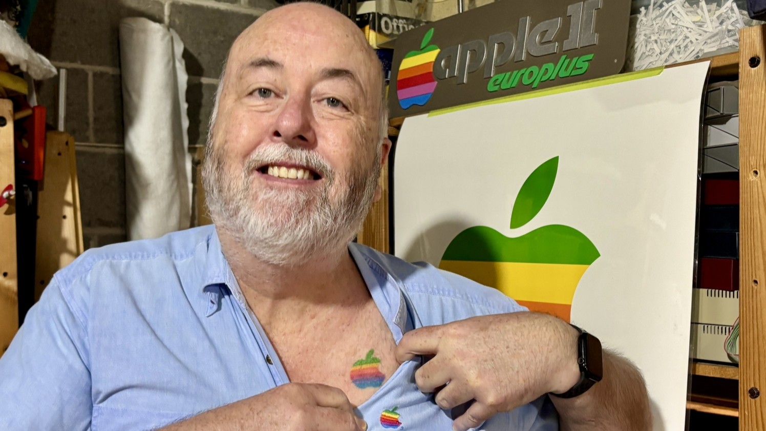 A close up of a middle-aged man wearing a shirt with an Apple logo, opening his shirt to reveal another Apple logo. Other Apple logos on the wall behind him.