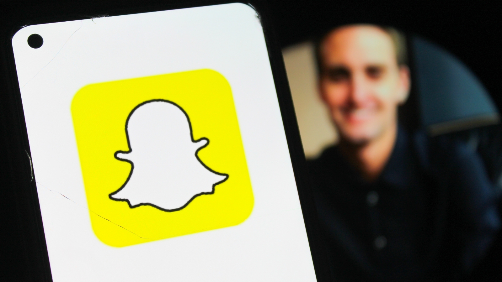 A close up of a smartphone displaying the Snapchat logo. In the background behind the phone, a blurred image of Evan Spiegel.