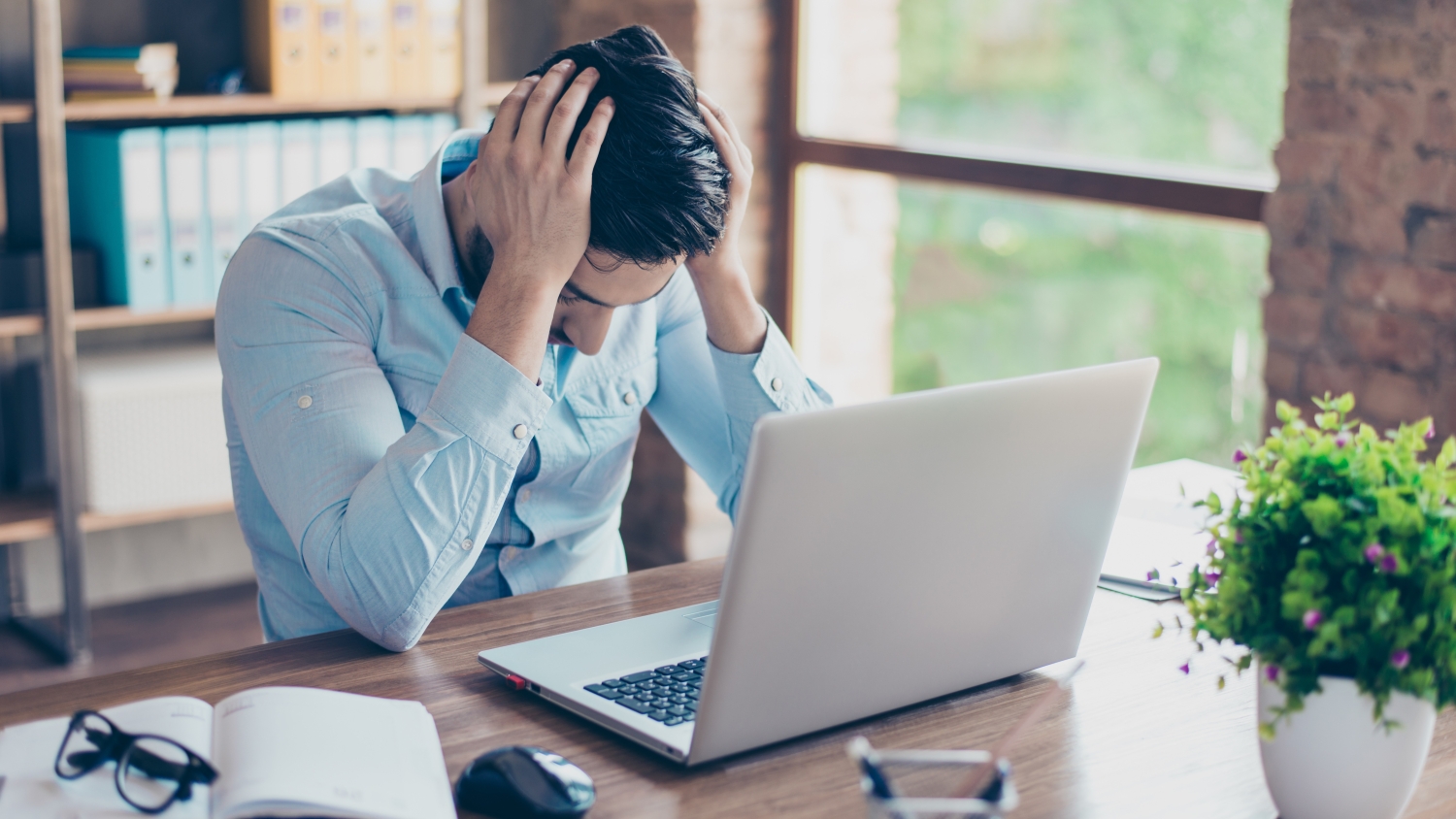A stressed male worker in an office holds his head in his hands and looks at his laptop