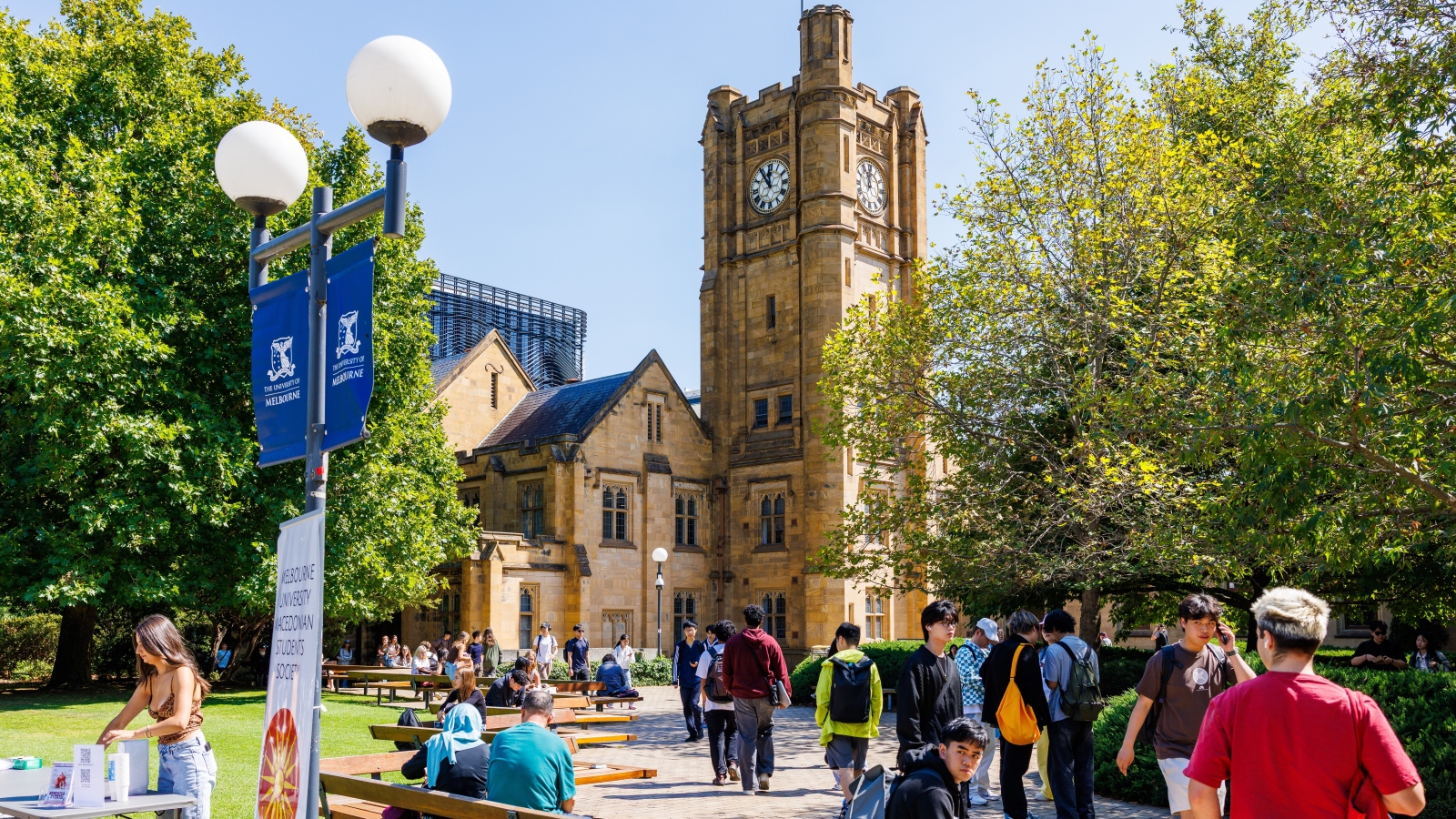 A wide outdoor shot of sandstone buildings, trees, and students at the University of Melbourne.