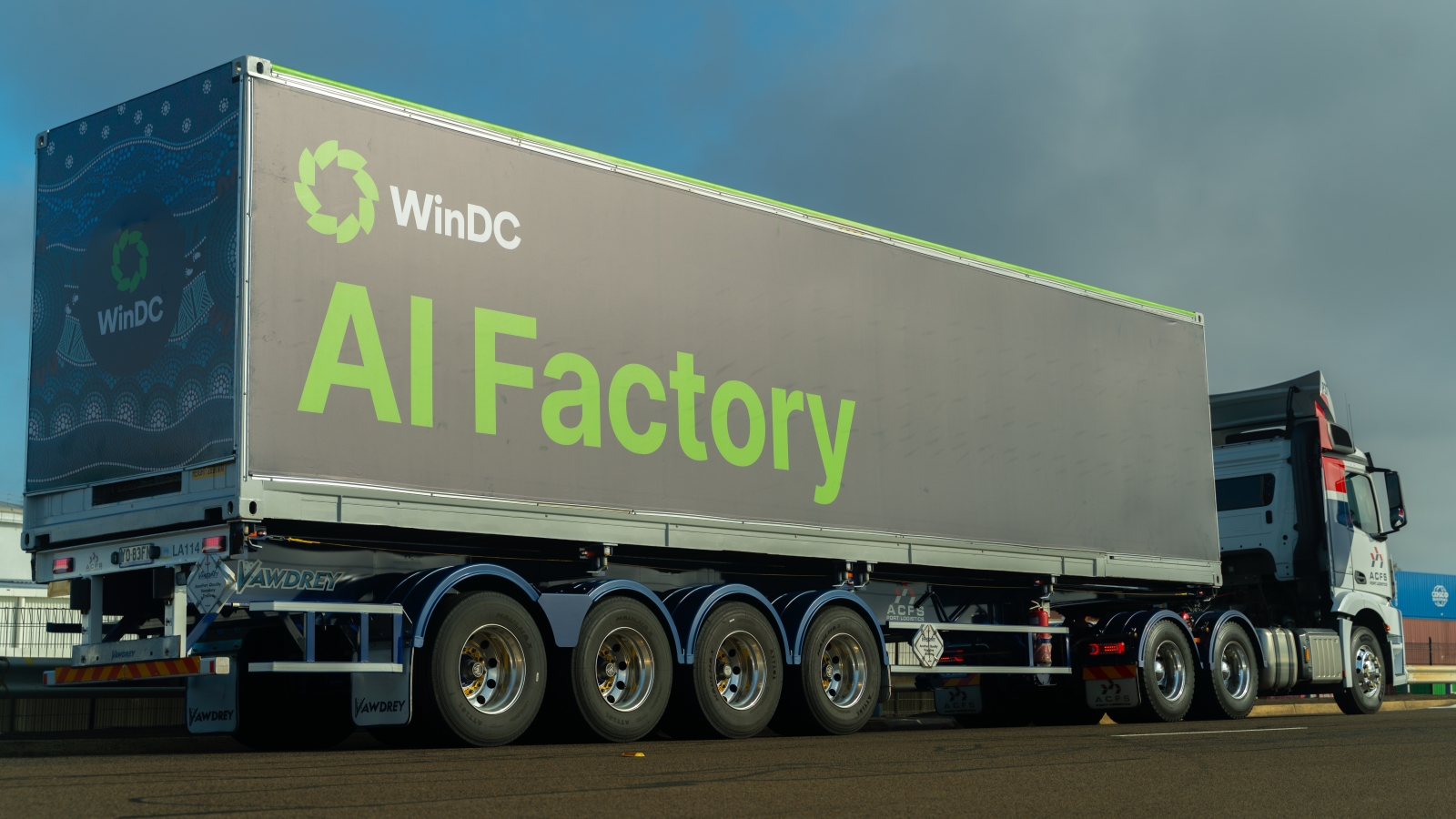 A wide outdoor shot of a large truck carrying a shipping container branded with the words 'WinDC AI Factory'