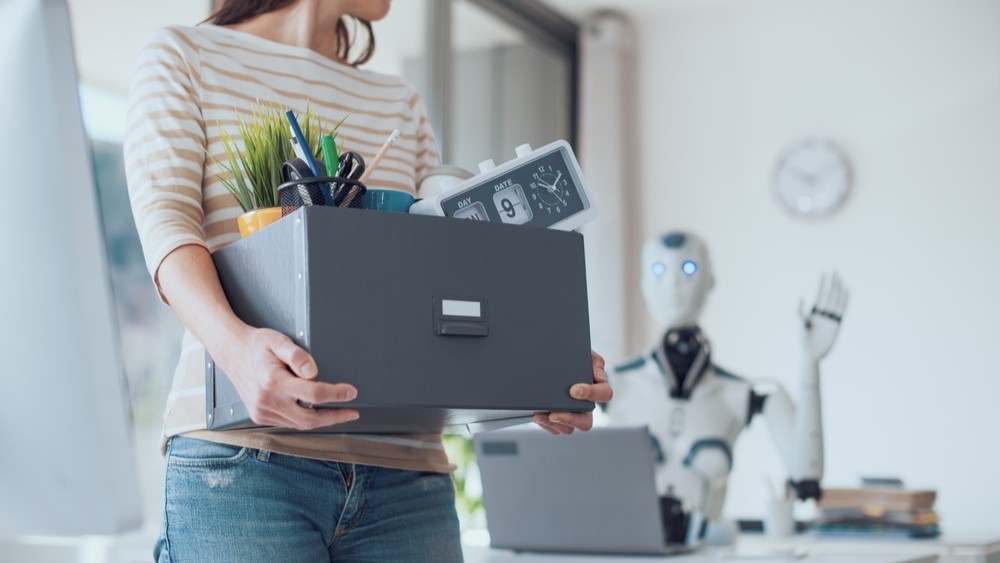 Photo of woman walking out of workplace holding box as AI robot waves goodbye.