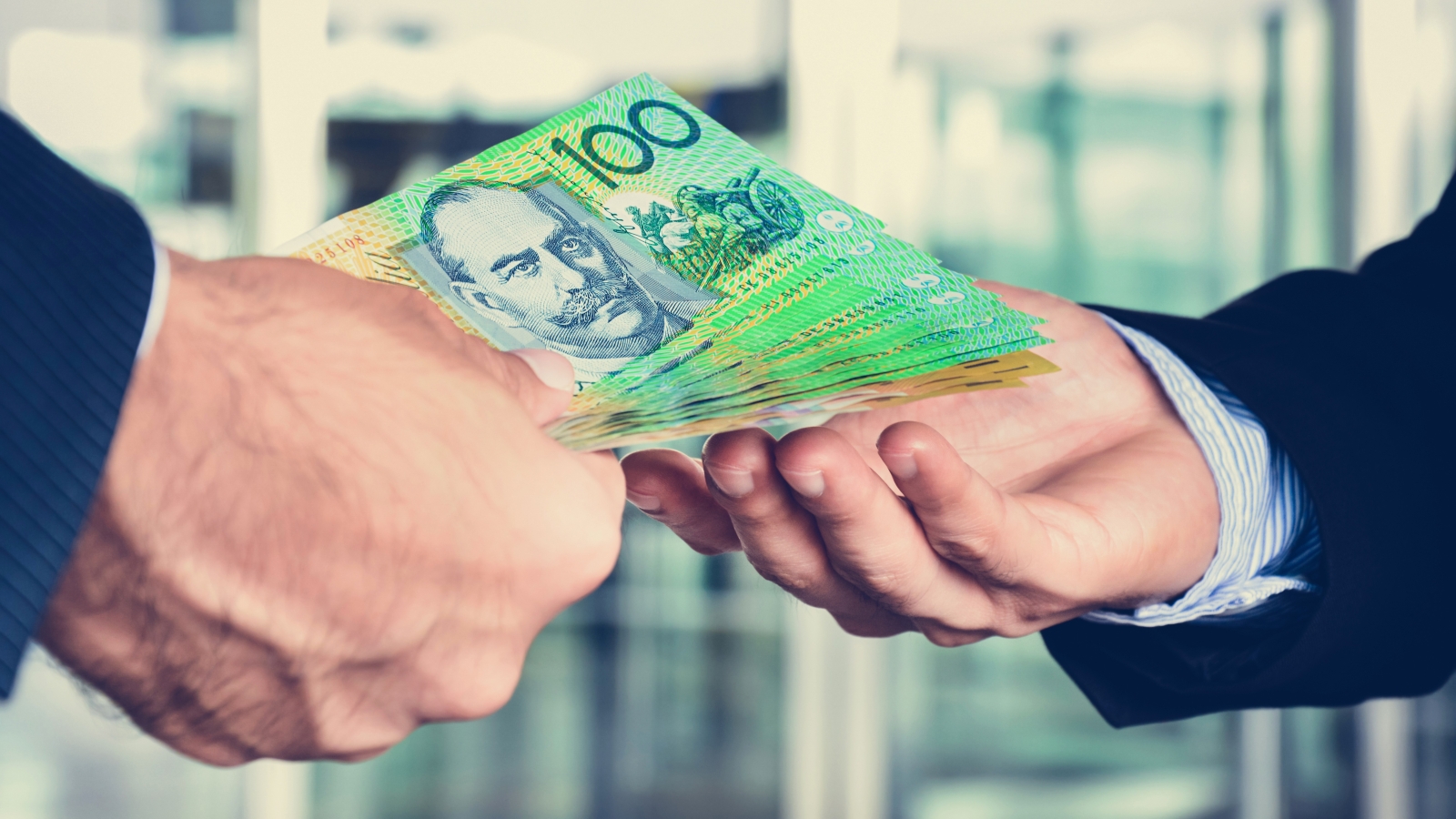 A close up of someone's hand passing a pile of $100 notes to another person's hand, inside an office.
