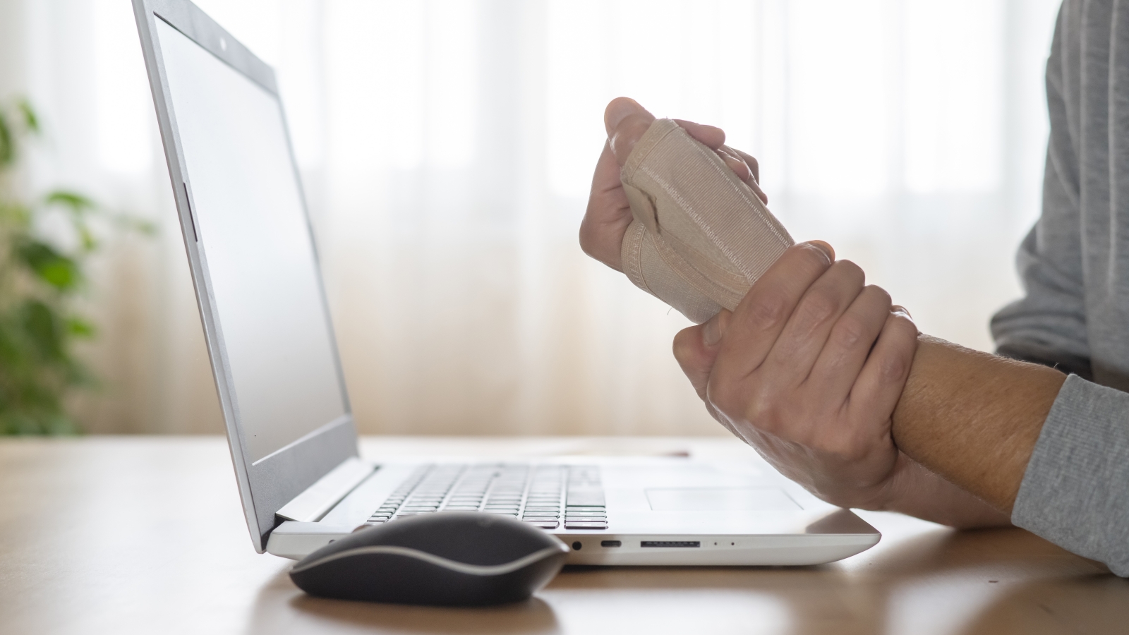 A close up of a person holding their injured wrist while working on a laptop and mouse.