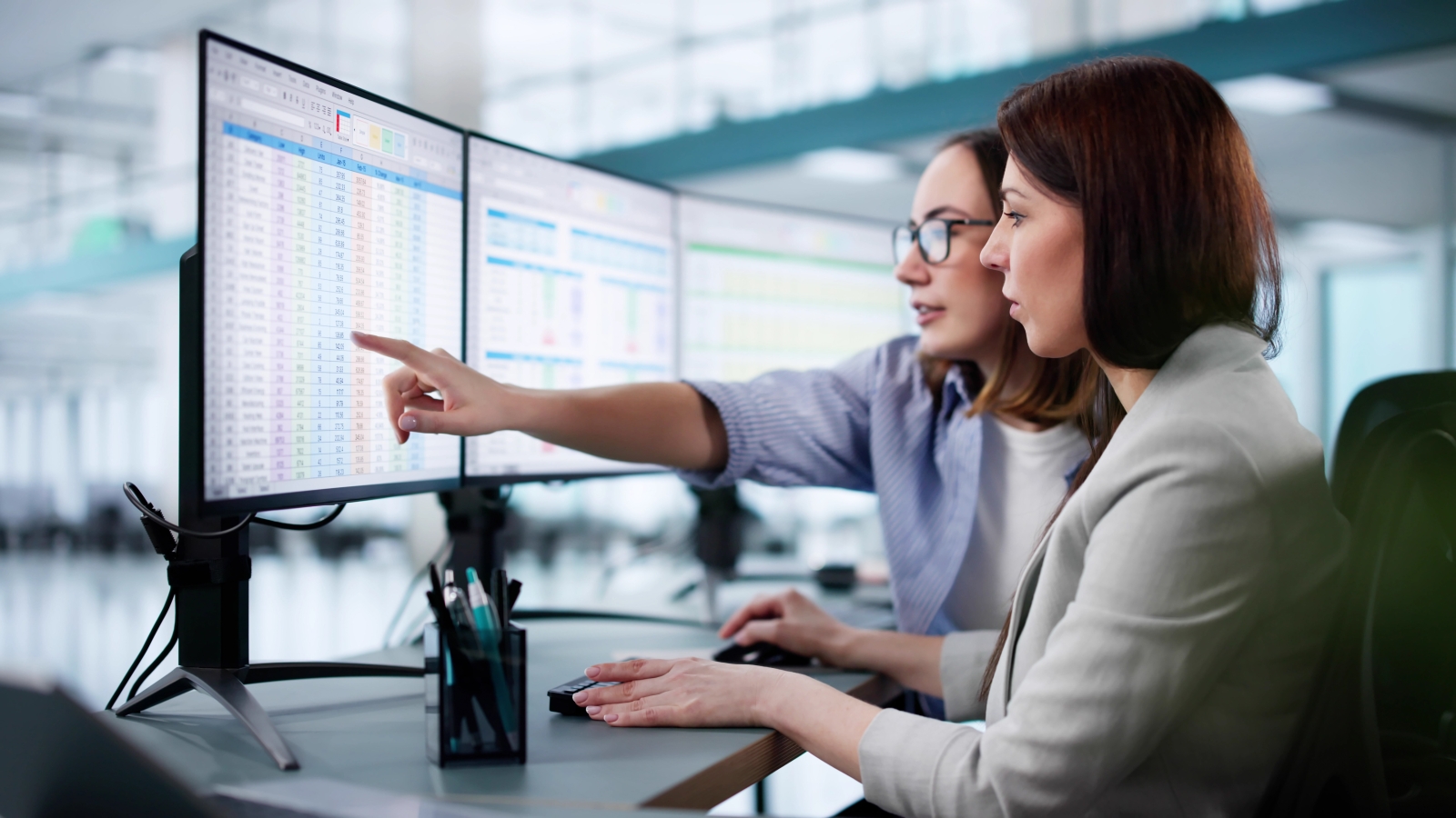 Two female workers examining large spreadsheets on computer screens in a modern office.
