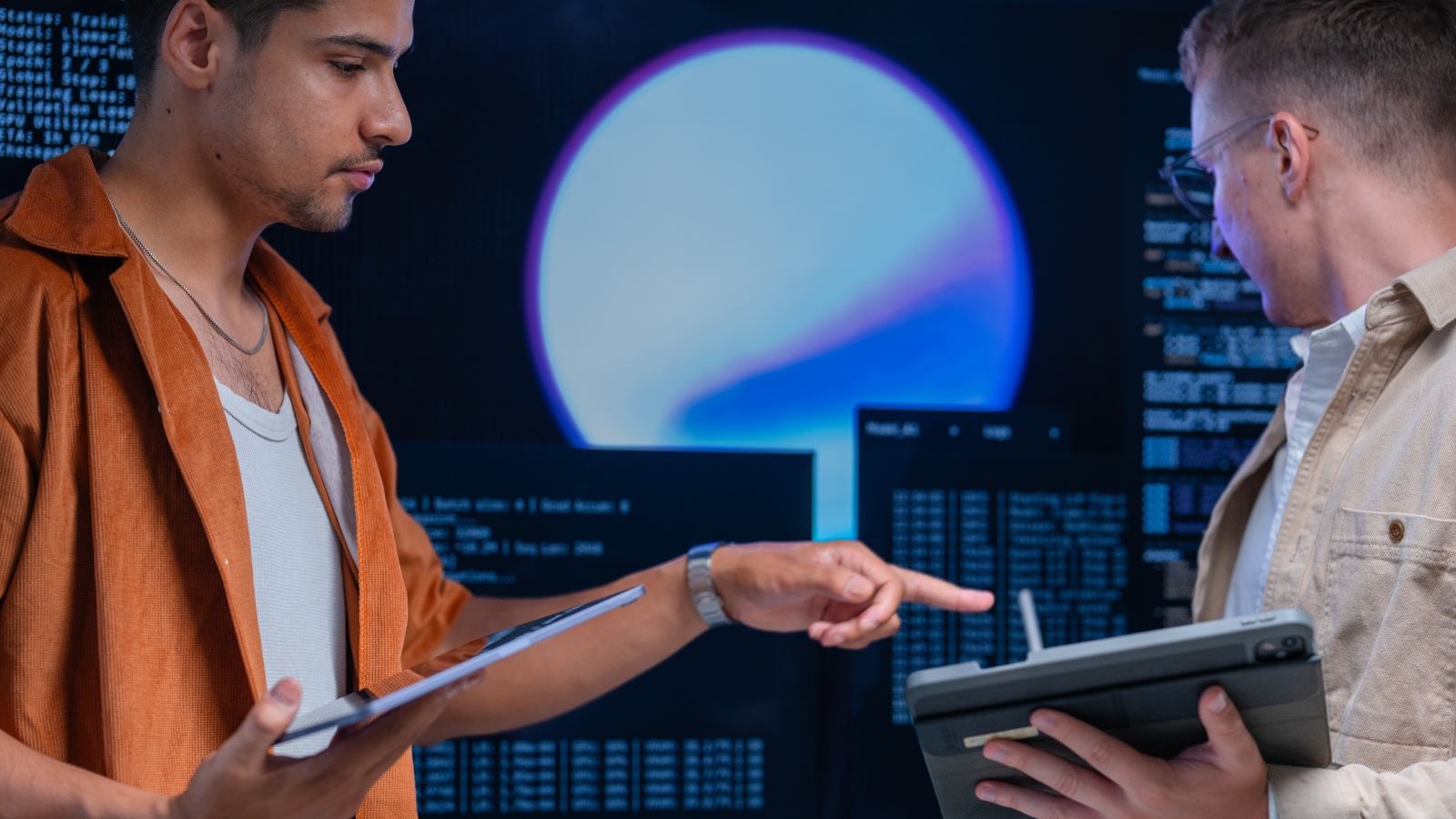 Two young male cybersecurity workers holding tablets and working in front of a screen displaying computer code.
