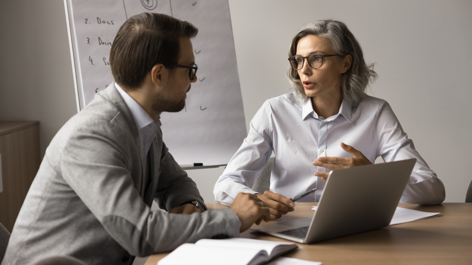  A medium shot of a younger male worker and an older female worker sitting at a desk in an office and chatting.